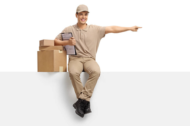 Delivery guy with packages and a clipboard seated on a panel pointing isolated on white background - Фото, зображення