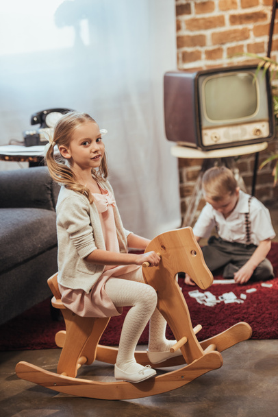 chica sentada en el caballo mecedora y mirando a la cámara y el hermano pequeño jugando con fichas de dominó detrás en casa, estilo de los años 50
 - Foto, Imagen