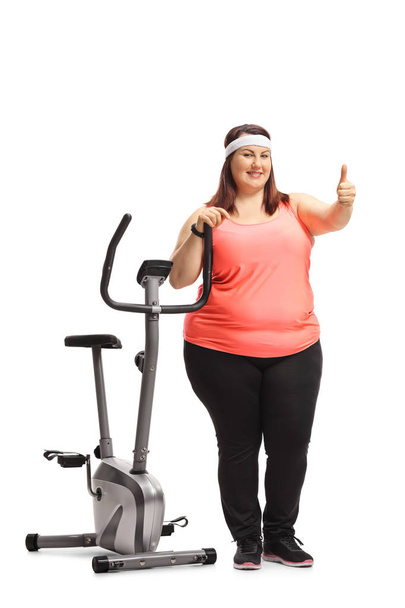 Full length portrait of an overweight woman leaning on an exercise bike and making a thumb up sign isolated on white background - Photo, Image