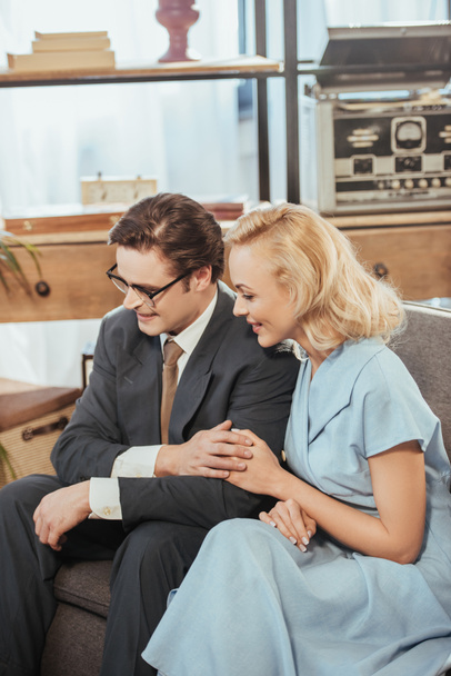 glückliches Paar im 50er-Jahre-Stil, das wegschaut, während es auf dem Sofa sitzt und Händchen hält - Foto, Bild