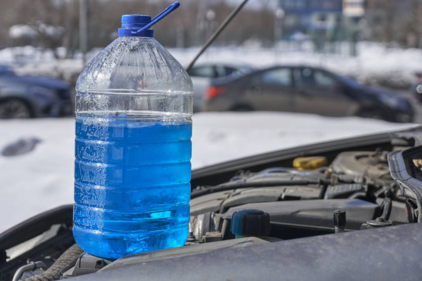 Caixote com líquido não congelante para lava pára-brisas em pé no motor de um carro em frente a uma estrada com carros no inverno ao ar livre
 - Foto, Imagem