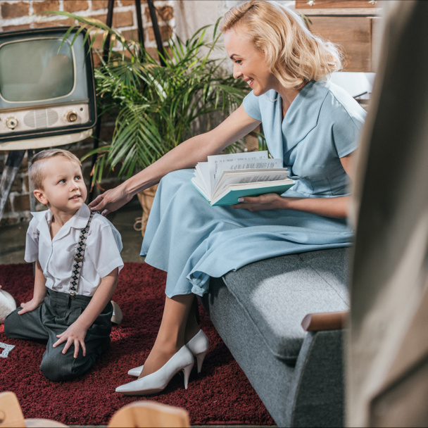 Gelukkig blonde vrouw boek lezen en kijken naar schattig zoontje spelen op tapijt thuis, jaren 1950 stijl familie - Foto, afbeelding