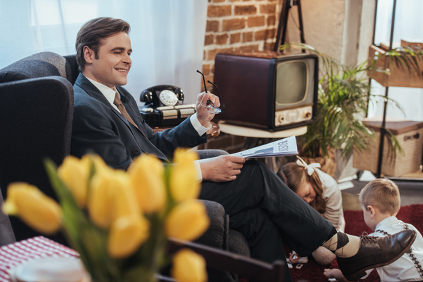 Lächelnder Mann im Anzug, der im Sessel Zeitung liest, während Kinder im Hintergrund mit Domino-Fliesen spielen, Familie im Stil der 1950er Jahre - Foto, Bild