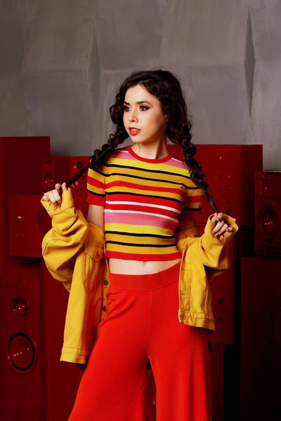 Studio fashion shooting. The girl in a bright outfit, top and pants flared in red and orange colors.Brunette with curly hair . Keeps your braids with your hands - Photo, image