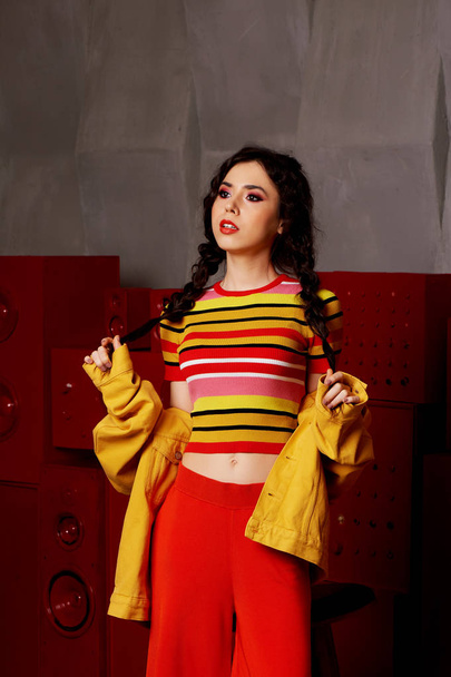 Studio fashion shooting. The girl in a bright outfit, top and pants flared in red and orange colors.Brunette with curly hair . Keeps your braids with your hands - Φωτογραφία, εικόνα
