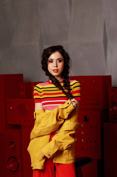 Studio fashion shooting. The girl in a bright outfit, top and pants flared in red and orange colors.Brunette with curly hair . Keeps your braids with your hands - 写真・画像