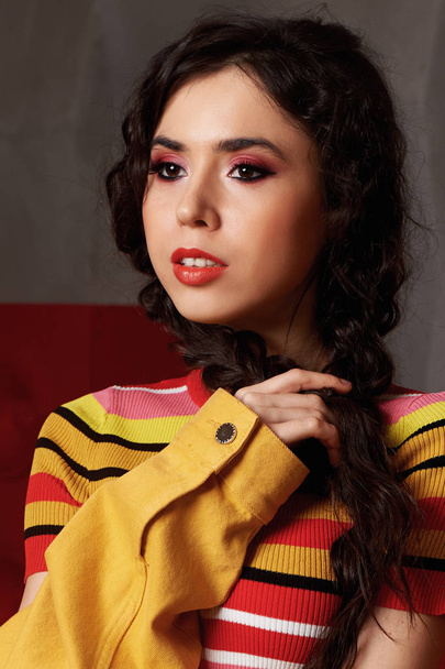 Studio fashion shooting. The girl in a bright outfit, top and pants flared in red and orange colors.Brunette with curly hair . Keeps your braids with your hands - Valokuva, kuva