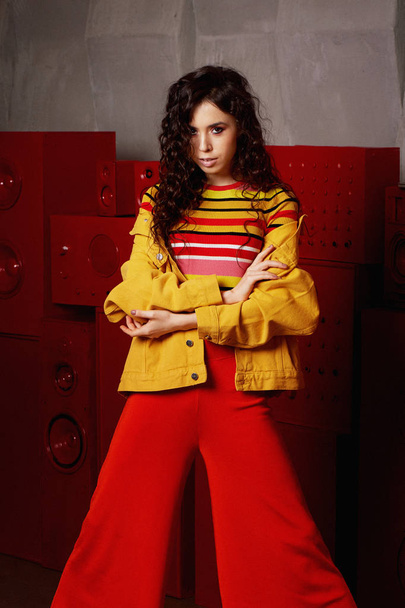 Studio fashion shooting.The girl in a bright outfit, top and pants flared in red and orange colors. Brunette with curly hair - Foto, Imagen
