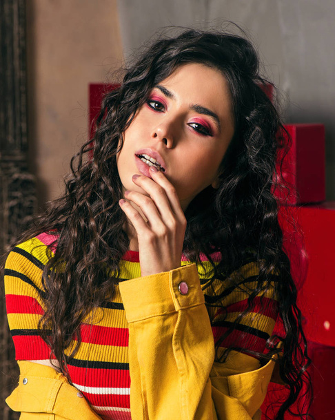 Studio fashion shooting. The girl in a bright outfit, top and pants flared in red and orange colors. Brunette with curly hair .Close-up portrait. Fingers around the lips - Zdjęcie, obraz