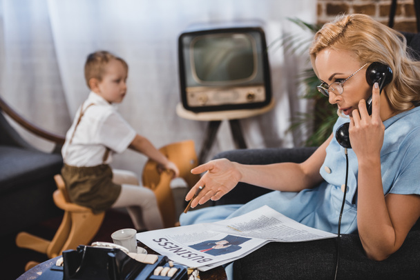 Frau mit Brille liest Wirtschaftszeitung und telefoniert mit Vintage-Telefon, während kleiner Sohn auf Schaukelpferd hinter der Familie im 50er-Jahre-Stil sitzt - Foto, Bild