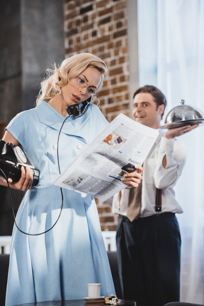 serious woman reading newspaper and talking by vintage telephone while husband holding meal behind, 50s style - Photo, Image
