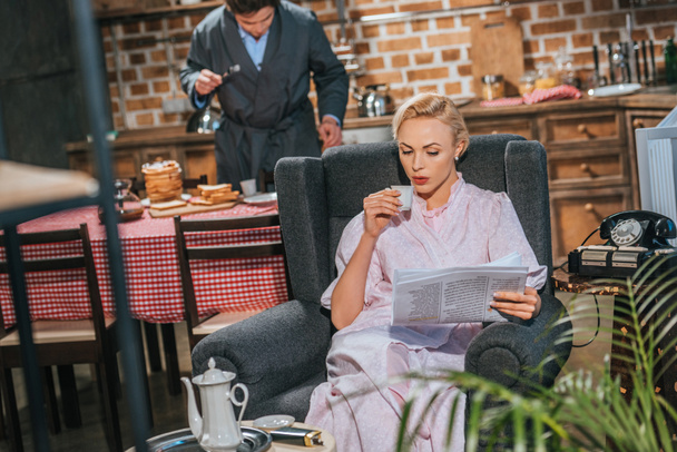 vrouw in gewaad drinken koffie en het lezen van de krant terwijl echtgenoot voorbereiding ontbijt achter, stijl van de jaren 1950 - Foto, afbeelding