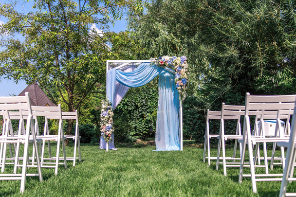 Arco de boda decorado con tela azul y flores al aire libre con filas de sillas blancas
 - Foto, imagen