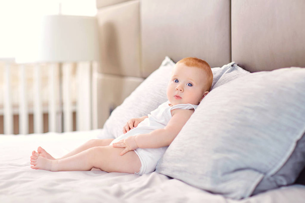 Funny baby is sitting on a pillow on the bed. - Zdjęcie, obraz