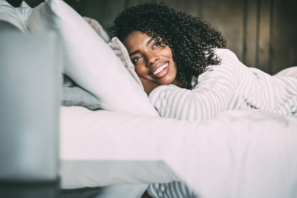 κοντινό πλάνο του μια όμορφη μαύρη γυναίκα με σγουρά μαλλιά χαμογελώντας και ξαπλωμένος στο κρεβάτι που βλέπει μακριά - Φωτογραφία, εικόνα