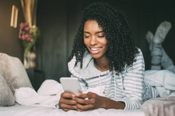 close up of a pretty black woman with curly hair smiling and using phone on bed looking away - Foto, Imagen