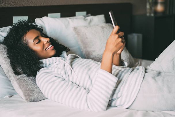zbliżenie na całkiem czarne kobiety z kręconymi włosami, uśmiechając się i przy użyciu telefonu na łóżku patrząc od hotelu - Zdjęcie, obraz