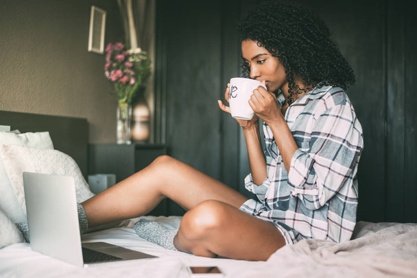bela mulher negra na cama com laptop e xícara de café
 - Foto, Imagem