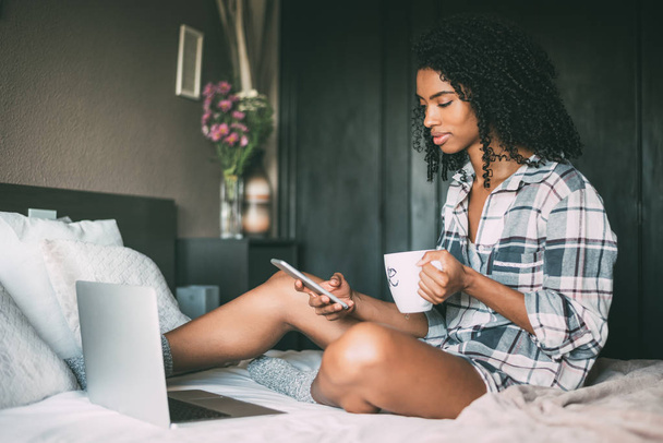 beautiful black woman on bed with laptop and cup of coffee - 写真・画像