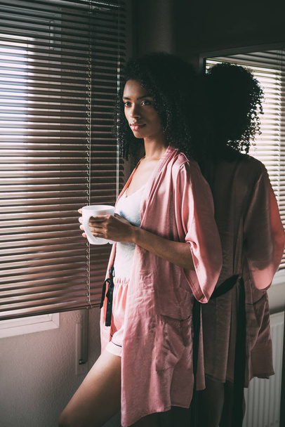 mooie zwarte vrouw stond bij het raam met kopje koffie - Foto, afbeelding