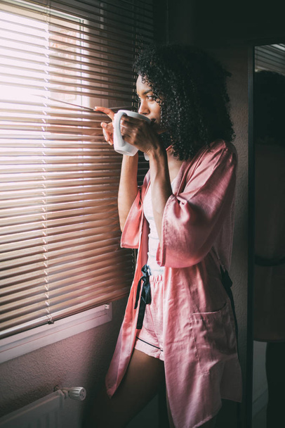 симпатичная чернокожая женщина, стоящая у окна с чашкой кофе
 - Фото, изображение