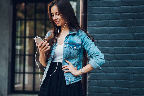 Elegáns pozitív blogger hallgat zenét a rádió honlapján online modern fülhallgató telefon csatlakozik. Vidám csípő lány beszélgető barátokkal, miközben kedvenc énekesmadár-ra smartphone - Fotó, kép
