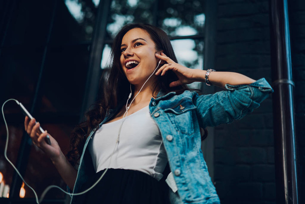 Below view of emotional positive brunette female holding modern smartphone in hand and listening favourite songs in earphones on radio website.Attractive young woman in denim jacket enjoying music - Foto, Imagem