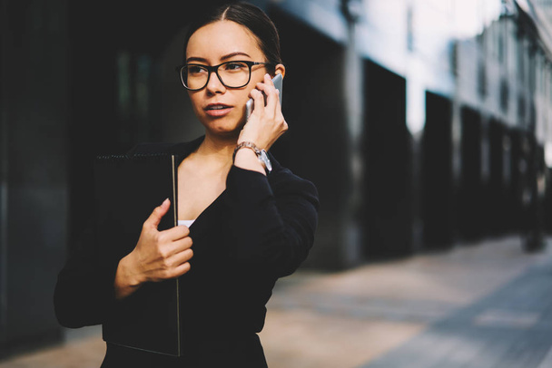 Avvocata giovane donna fiduciosa in occhiali ottici che parla con i partner commerciali tramite smartphone moderno mentre si trova all'aperto e tiene in mano la cartella.
 - Foto, immagini
