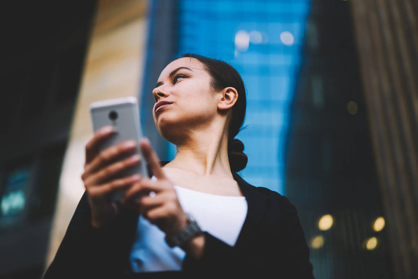 Below view of confident professional lawyer sending sms message on digital smartphone while looking away standing outdoors on office promotional background.Serious female banker holding cellular - Photo, image