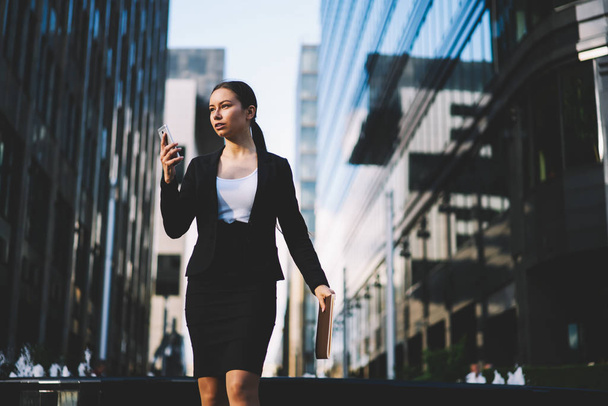 Confident businesswoman dressed in black formal apparel sending sms message to colleague on digital smartphone and holding folder in hand while strolling on office building promotional background - Photo, image