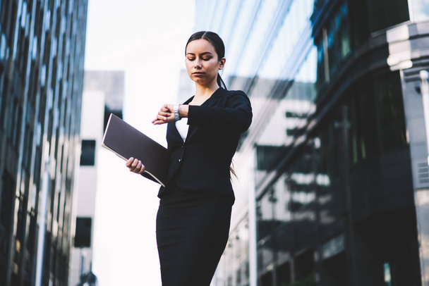 Serious businesswoman holding folder in hand and looking at wristwatch to checking time standing outdoors near office building.Confident female banker in formal wear tracking clock on smartwatch - Foto, imagen