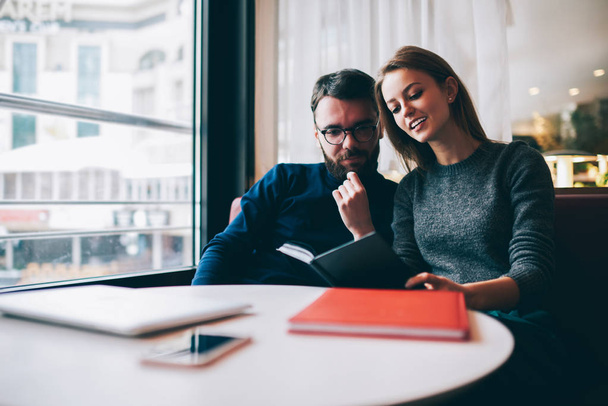 Friends sitting in comfortable coffee shop and reading book.Bearded man in eyeglasses and attractive woman studying together in coworking space.Male and female hipsters discussing text information - Photo, Image