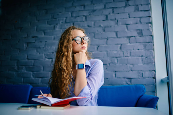 Thoughtful young woman with blonde curly hair and optical eyeglasses sitting with books in coworking space and looking out of window.Contemplative student in spectacles thinking on ideas - Foto, afbeelding