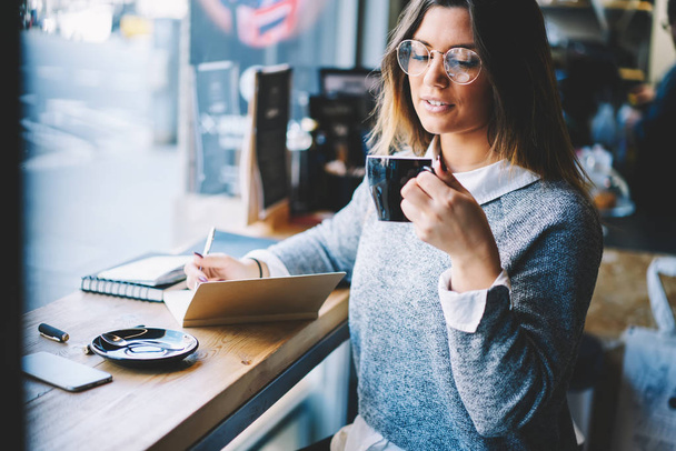 Successful female owner in optical eyeglasses enjoying cappuccino while noting plans in notebook sitting at wooden table in coffee shop interior. - Fotoğraf, Görsel