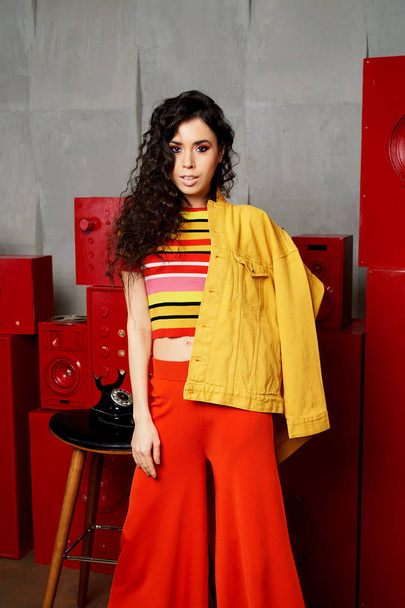 Studio fashion shooting. The girl in a bright outfit, top and pants flared in red and orange tones. Brunette with curly hair. Beautiful and unusual pose. Attractive - Foto, Imagem