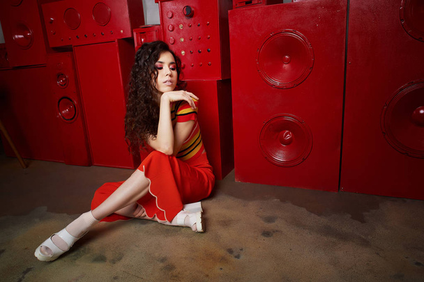 Studio fashion shooting. The girl in a bright outfit, top and pants flared in red and orange tones. Brunette with curly hair. Beautiful and unusual pose. Attractive - Φωτογραφία, εικόνα