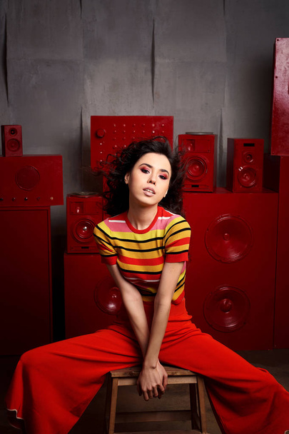Studio fashion shooting. The girl in a bright outfit, top and pants flared in red and orange tones. Brunette with curly hair. Beautiful and unusual pose. Attractive - 写真・画像