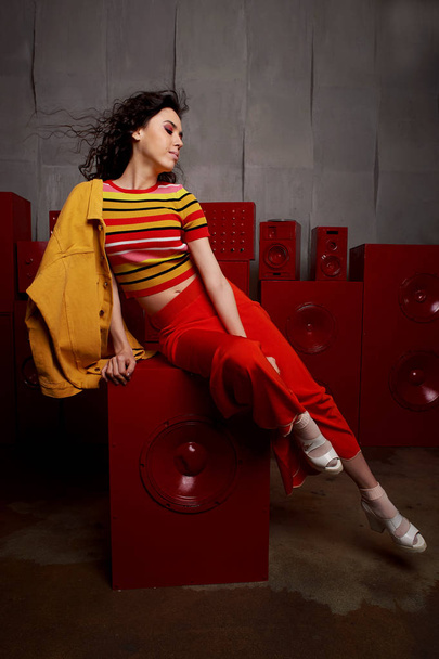 Studio fashion shooting. The girl in a bright outfit, top and pants flared in red and orange tones. Brunette with curly hair. Beautiful and unusual pose. Attractive - Foto, Bild
