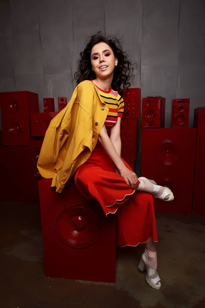 Studio fashion shooting. The girl in a bright outfit, top and pants flared in red and orange tones. Brunette with curly hair. Beautiful and unusual pose. Attractive - Foto, afbeelding
