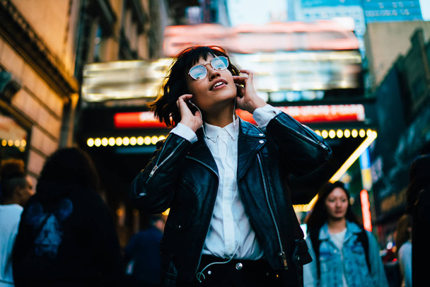 Positive hipster girl in trendy clothes listening favorite song from playlist while strolling on crowded city street in evening, young woman enjoying sound in earphones walking at night downtown - Foto, imagen
