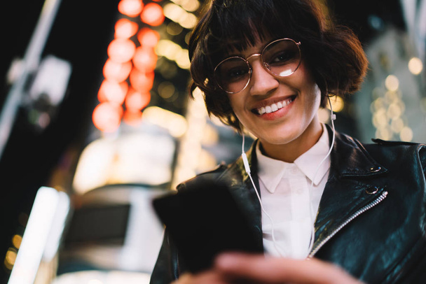 Portrait of cheerful hipster girl using application on smartphone enjoying nightlife in city, cheerful young woman sending message on smartphone standing near copy space - Φωτογραφία, εικόνα