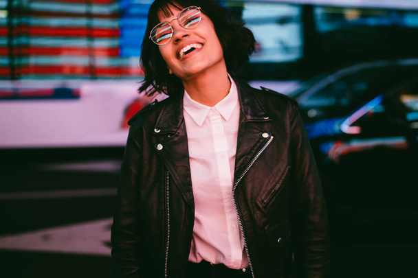 Cheerful hipster girl in spectacles with night city light reflection laughing feeling carefree on free time, smiling young woman in stylish clothing having fun in evening spending time in megalopoli - Фото, изображение