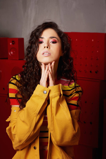 Studio fashion shooting.The girl in a bright outfit, top and pants flared in red and orange tones. Brunette with curly hair.Touching hands to the chin. - Foto, imagen