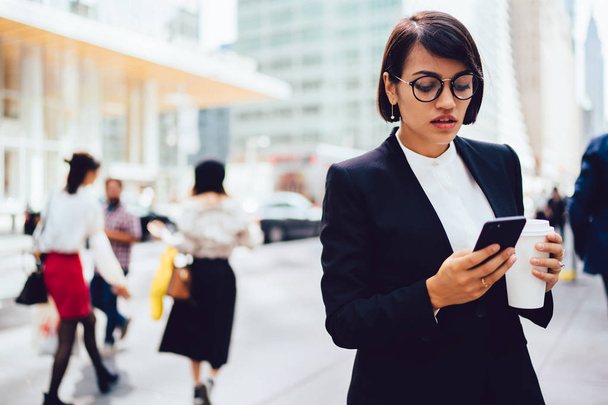 Serious female manager in formal wear checking notification on smartphone holding coffee to go, confident businesswoman in stylish spectacles browsing webpage for bank for checking account - Fotografie, Obrázek