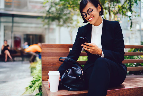 Smiling businesswoman in elegant clothes enjoying rest of coffee break sitting outdoors on bench chatting in social networks on mobile. - Photo, image