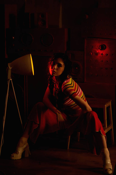 Studio fashion shooting.The girl in a bright outfit, top and pants flared in red and orange colors. Brunette with curly hair. Red light lamp in the twilight. The atmosphere of mystery - Zdjęcie, obraz