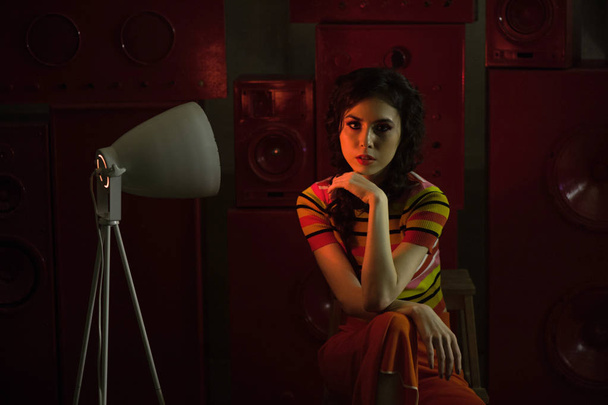 Studio fashion shooting.The girl in a bright outfit, top and pants flared in red and orange colors. Brunette with curly hair. Red light lamp in the twilight. The atmosphere of mystery - Foto, Imagem