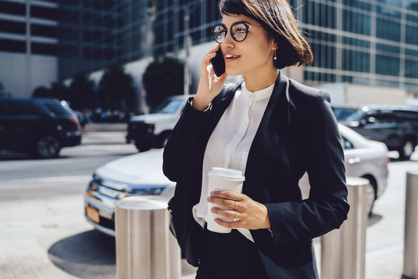Successful female manager in elegant wear walking on city street and talking on mobile, prosperous businesswoman having phone conversation about work holding coffee to go with copy space for brand nam - Fotografie, Obrázek