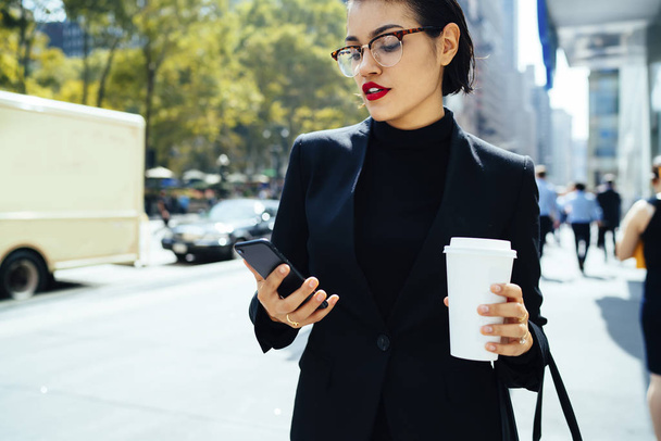 Confident brunette businesswoman reading income message on mobile while walking on street, serious female entrepreneur in elegant outfit and red lips checking email on phone strolling with coffee - Foto, Imagen
