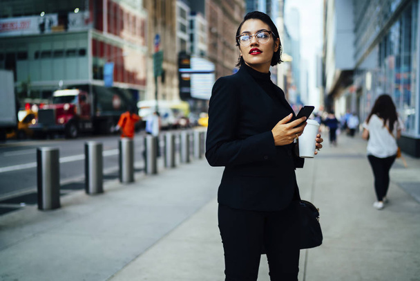 Trendily dressed female manager holding smartphone checking mail while strolling on manhattan street,beautiful businesswoman with red lips and in eyeglasses walking with coffee to go sending messages - Fotografie, Obrázek